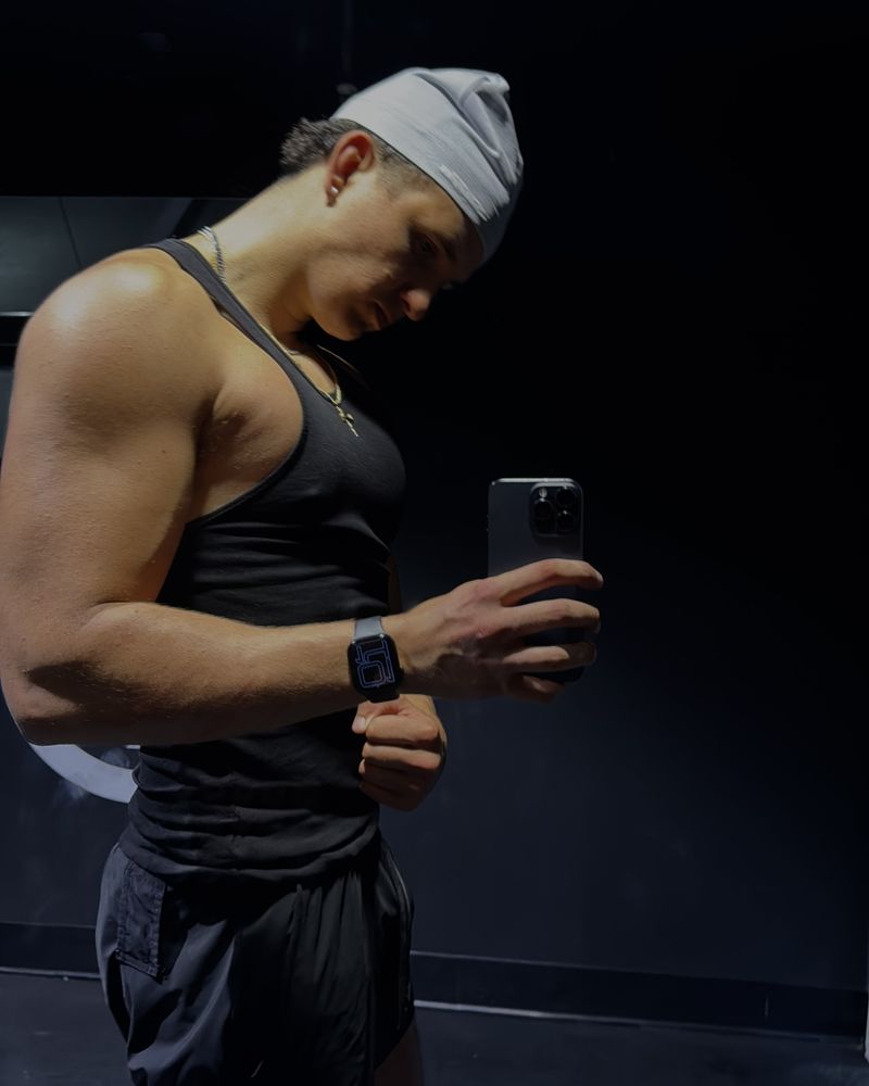Adam in a black tank top, flexing his arm in a gym mirror